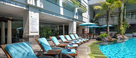 A poolside area with lounge chairs and umbrellas, a clear blue pool, and a building with balconies.