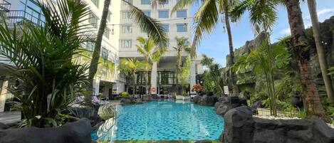 A pool area with a clear blue pool, surrounded by palm trees and rocks, with a building in the background.