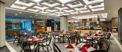 A spacious dining area with multiple tables and chairs, a modern ceiling design, and a view of the kitchen area in the background.
