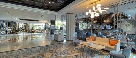 Un hall amplio con diseño moderno, que presenta una alfombra estampada, zona de estar con sofás y sillones, y una escalera con barandilla de metal.