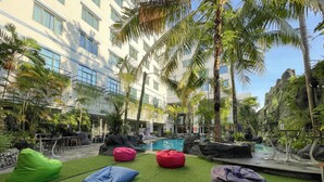 Zona junto a la piscina con césped artificial, puffs de colores y vista a un edificio con balcones y piscina.
