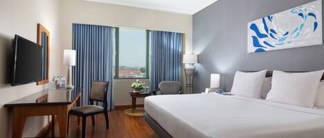 Habitación de hotel con cama, escritorio, silla, televisor, lámpara y ventana con cortinas.