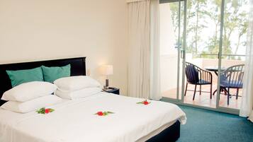 A hotel room with a bed, a nightstand, a lamp, a balcony with chairs, and a view of trees.