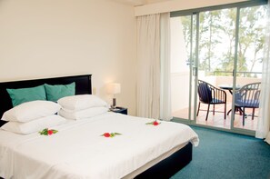 Een hotelkamer met een bed, nachtkastje, lamp, balkon met stoelen en uitzicht op bomen.