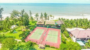A tennis court surrounded by lush greenery and a beach view.