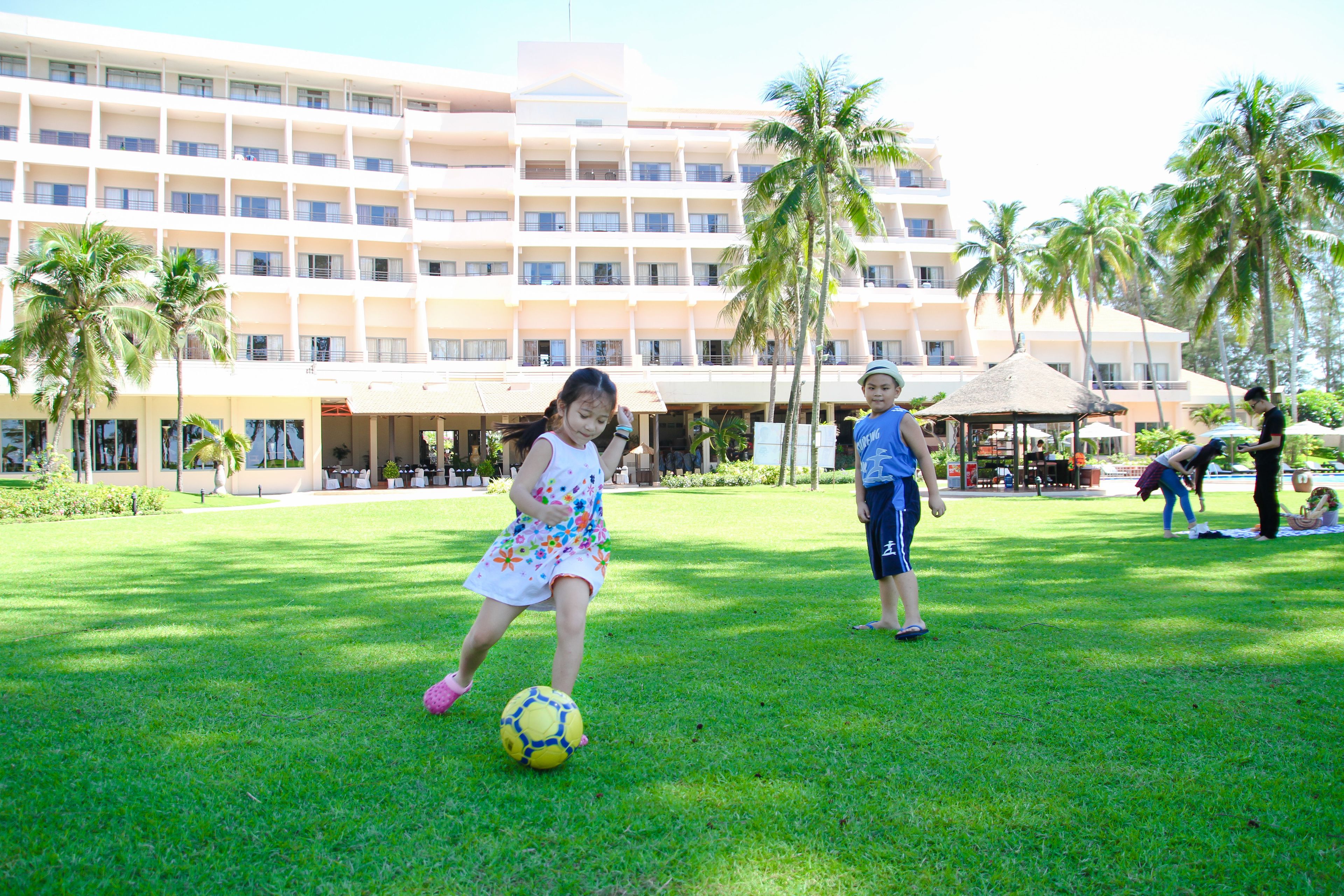 Un hotel con un edificio grande, palmeras y un área verde donde juegan niños.