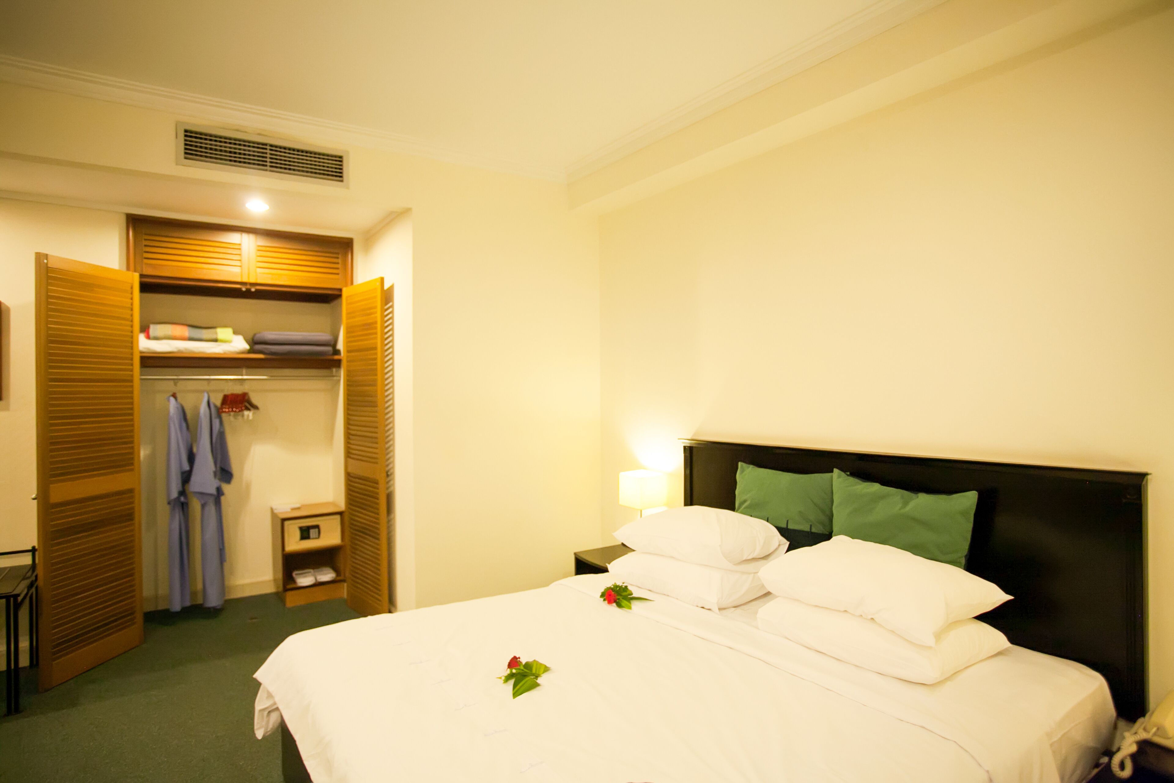 A hotel room with a neatly made bed, a bedside table with a lamp, a wardrobe, and a small shelf with folded towels.