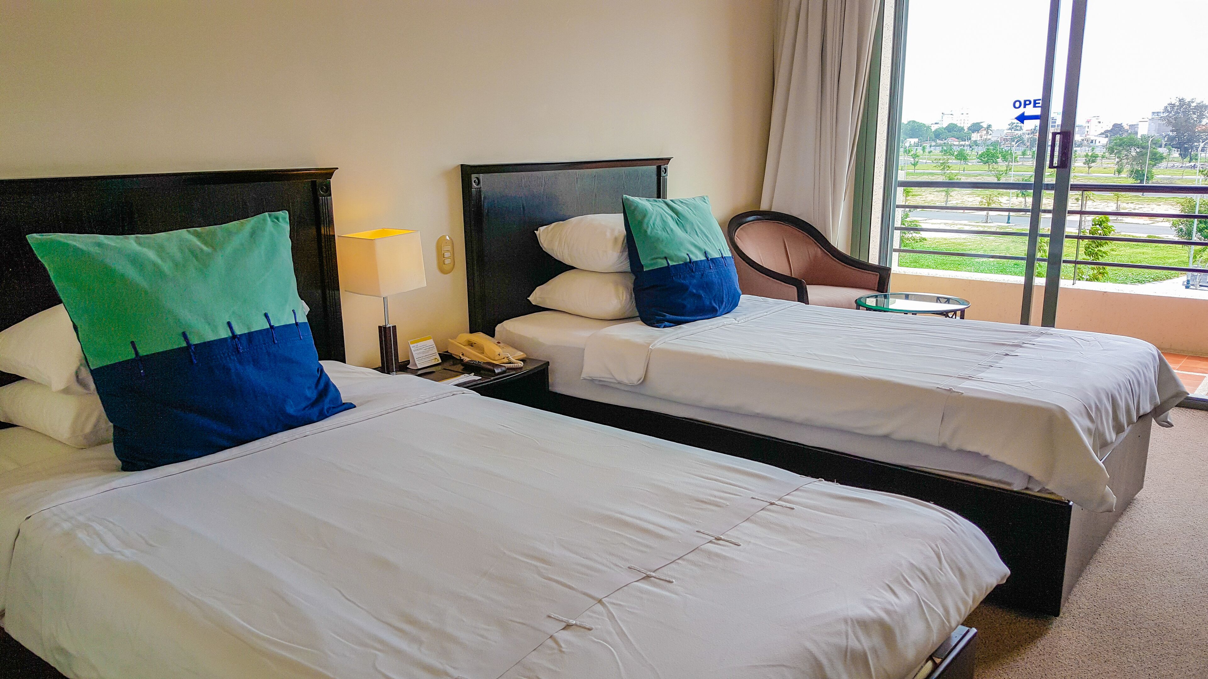 Two single beds with white bedding and green and blue pillows, a bedside table with a lamp, and a view of a cityscape through a large window.