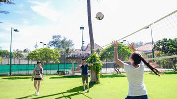 A volleyball game on a green artificial turf court with a fence and palm trees in the background.