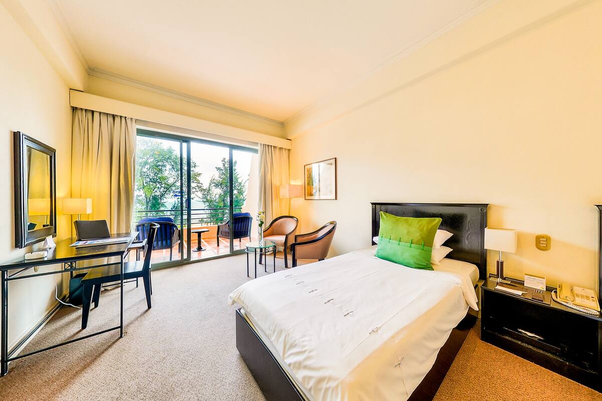 Une chambre d’hôtel comprenant un lit, un bureau avec un ordinateur portable, une chaise, une petite table avec un vase et une vue sur l’extérieur grâce à de grandes fenêtres.
