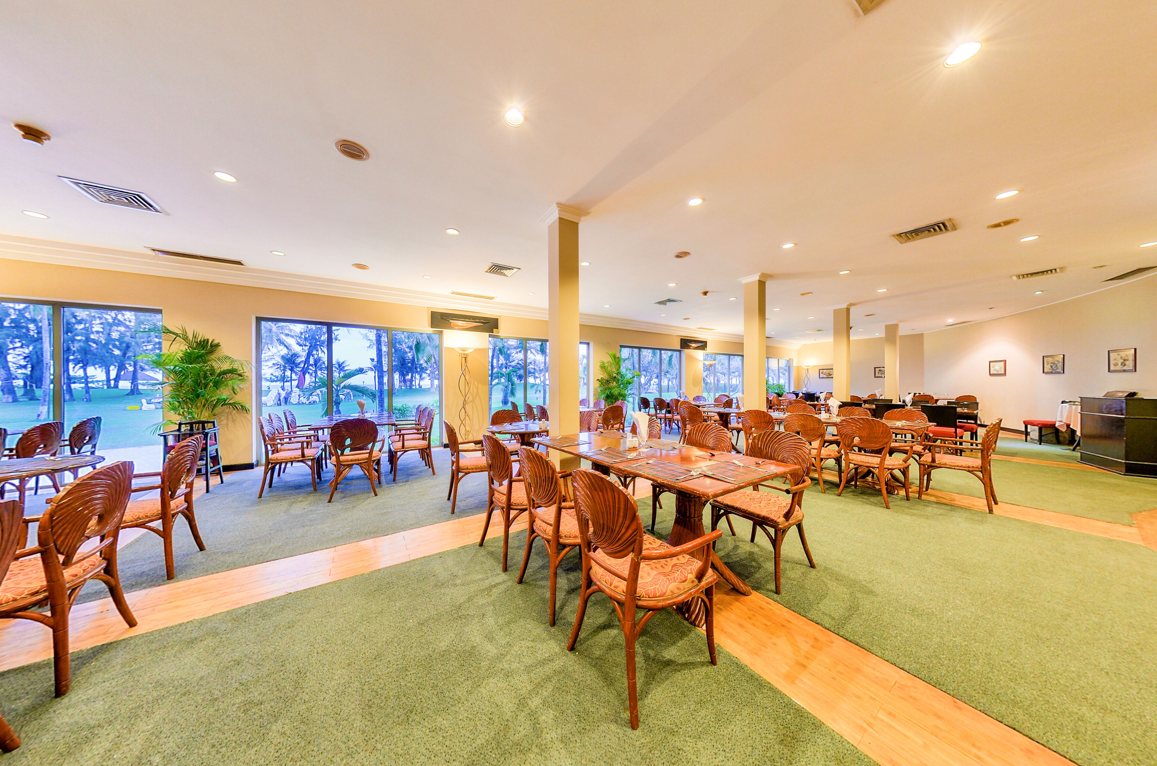 A spacious indoor dining area with multiple tables and chairs, large windows offering views of palm trees, and a green carpeted floor.