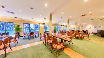 A spacious indoor dining area with multiple tables and chairs, large windows offering views of palm trees, and a green carpeted floor.