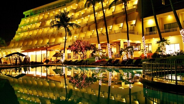 A multi-story hotel with a pool reflecting the building and palm trees.