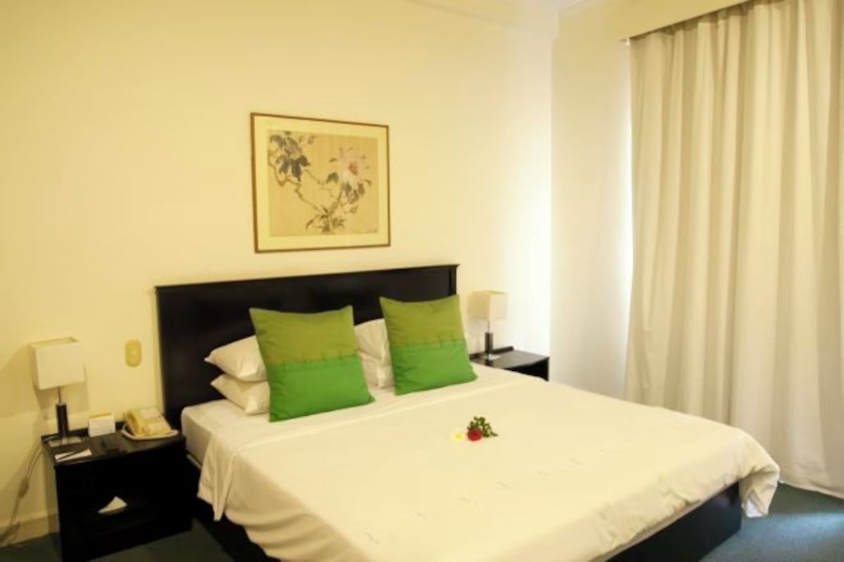 A hotel room with a bed, green pillows, a nightstand with a lamp, and a framed picture on the wall.