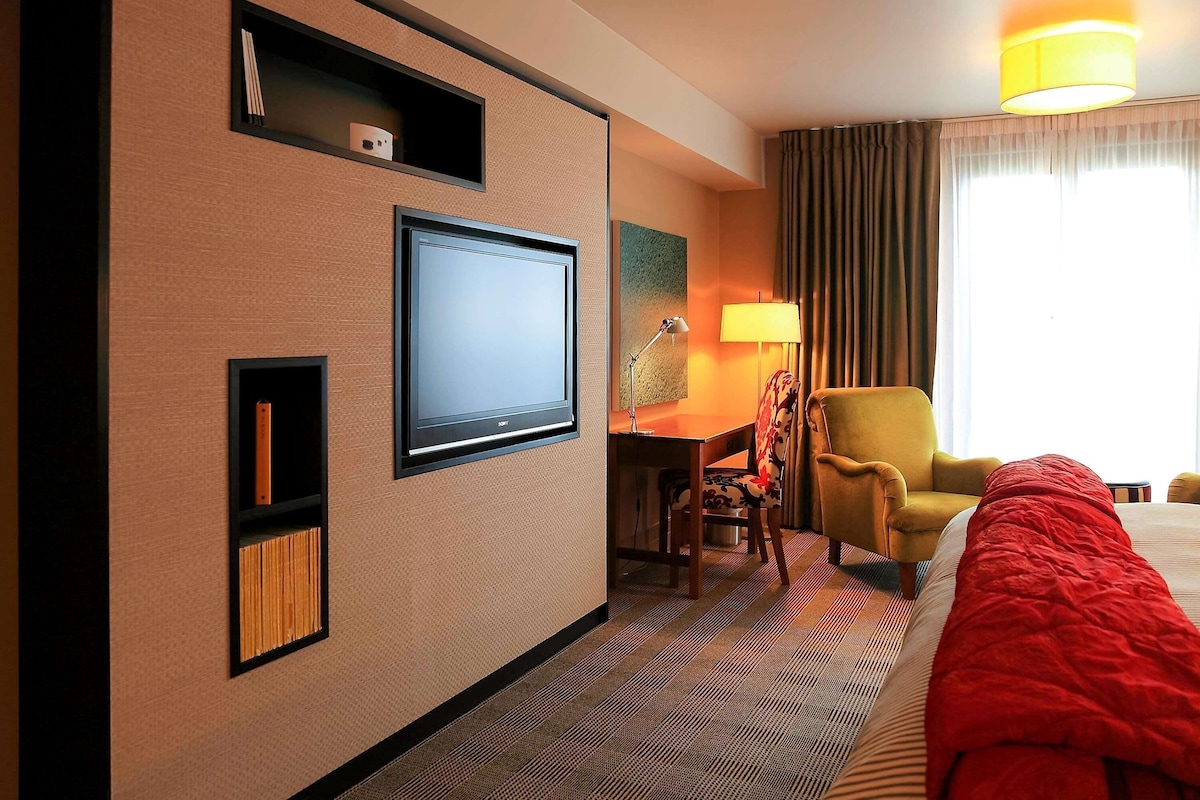 A hotel room with a flat-screen TV, a desk with a lamp, a yellow armchair, a red blanket, and a large window with curtains.