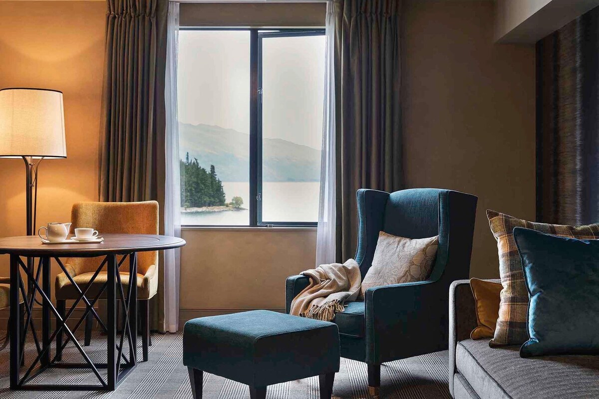 A hotel room with a view of a lake and mountains, a blue armchair, a sofa, a small table with a lamp, and a chair.