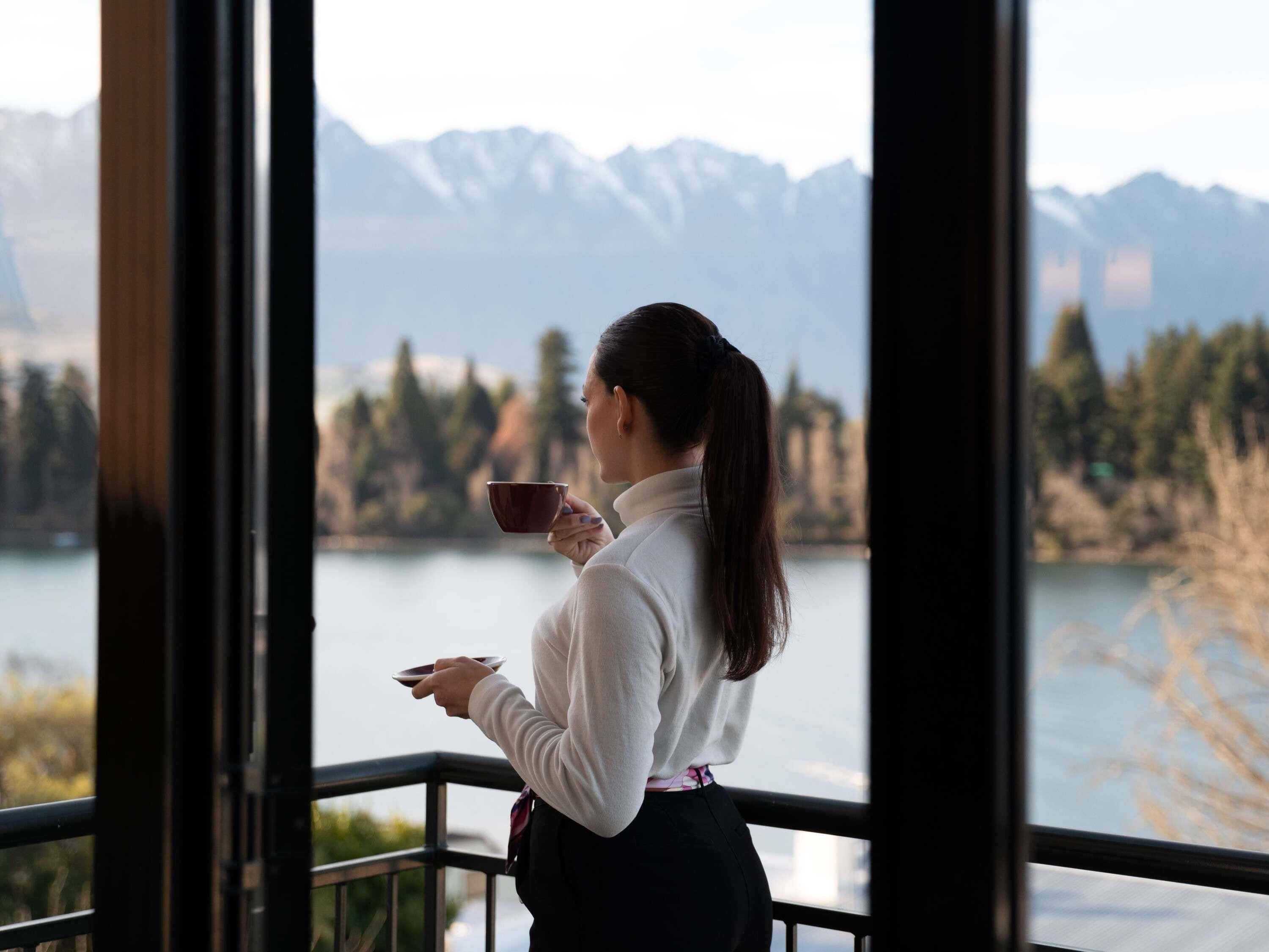A person holding a cup, standing by a window with a view of mountains and trees.