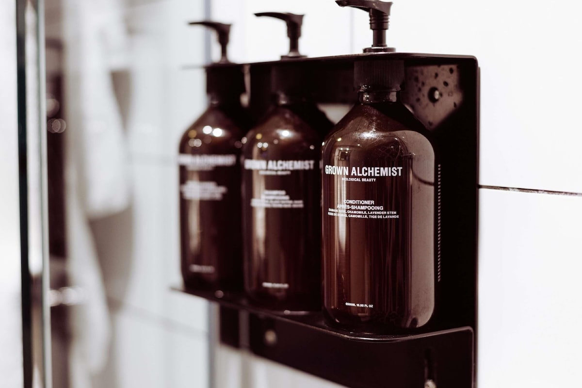 A row of three dark-colored bottles with pump dispensers, labeled 'GROWN ALCHEMIST,' on a black shelf.