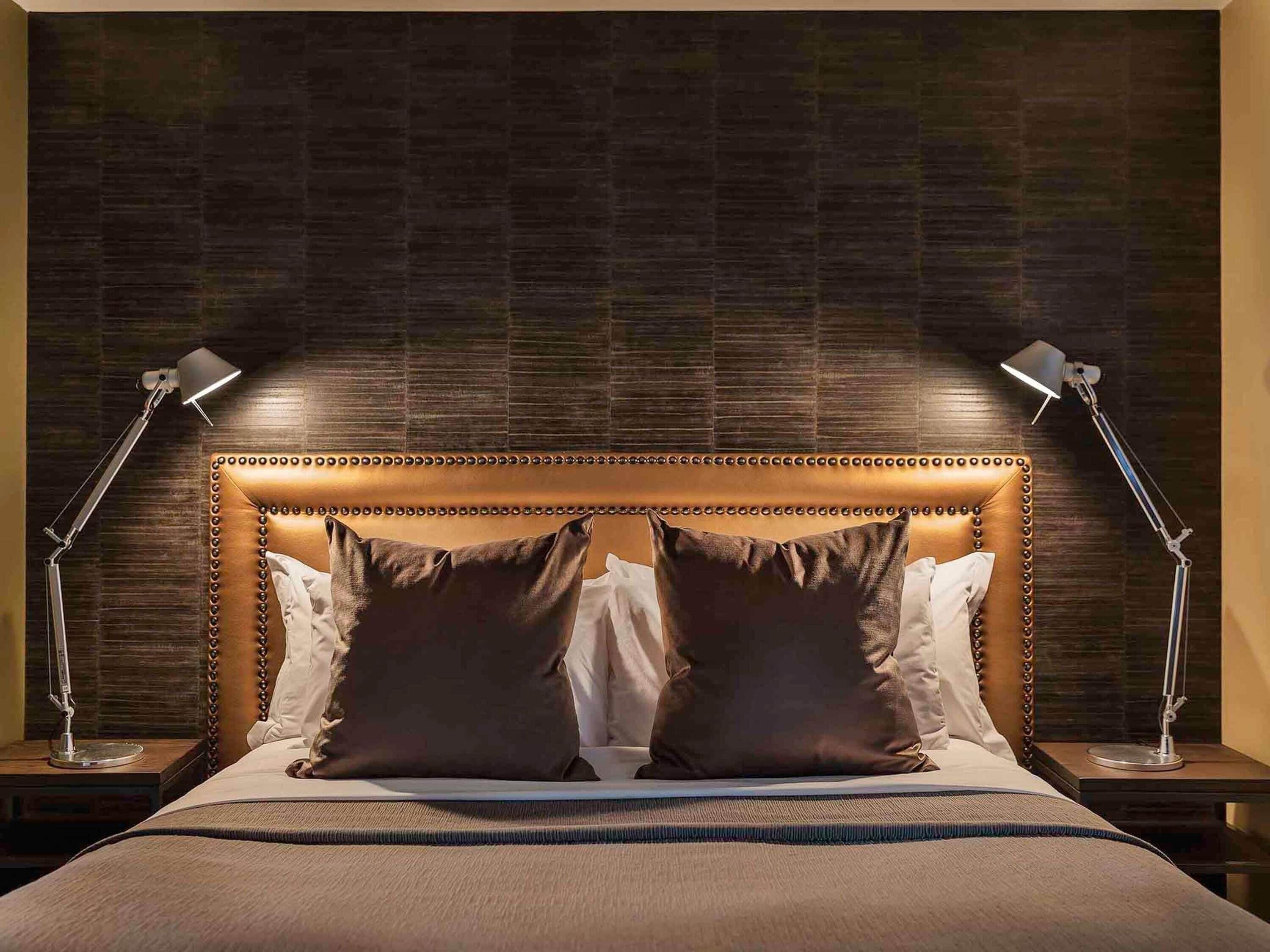 Uma cama com cabeceira estofada, duas luminárias de apoio e uma parede com padrão escuro.