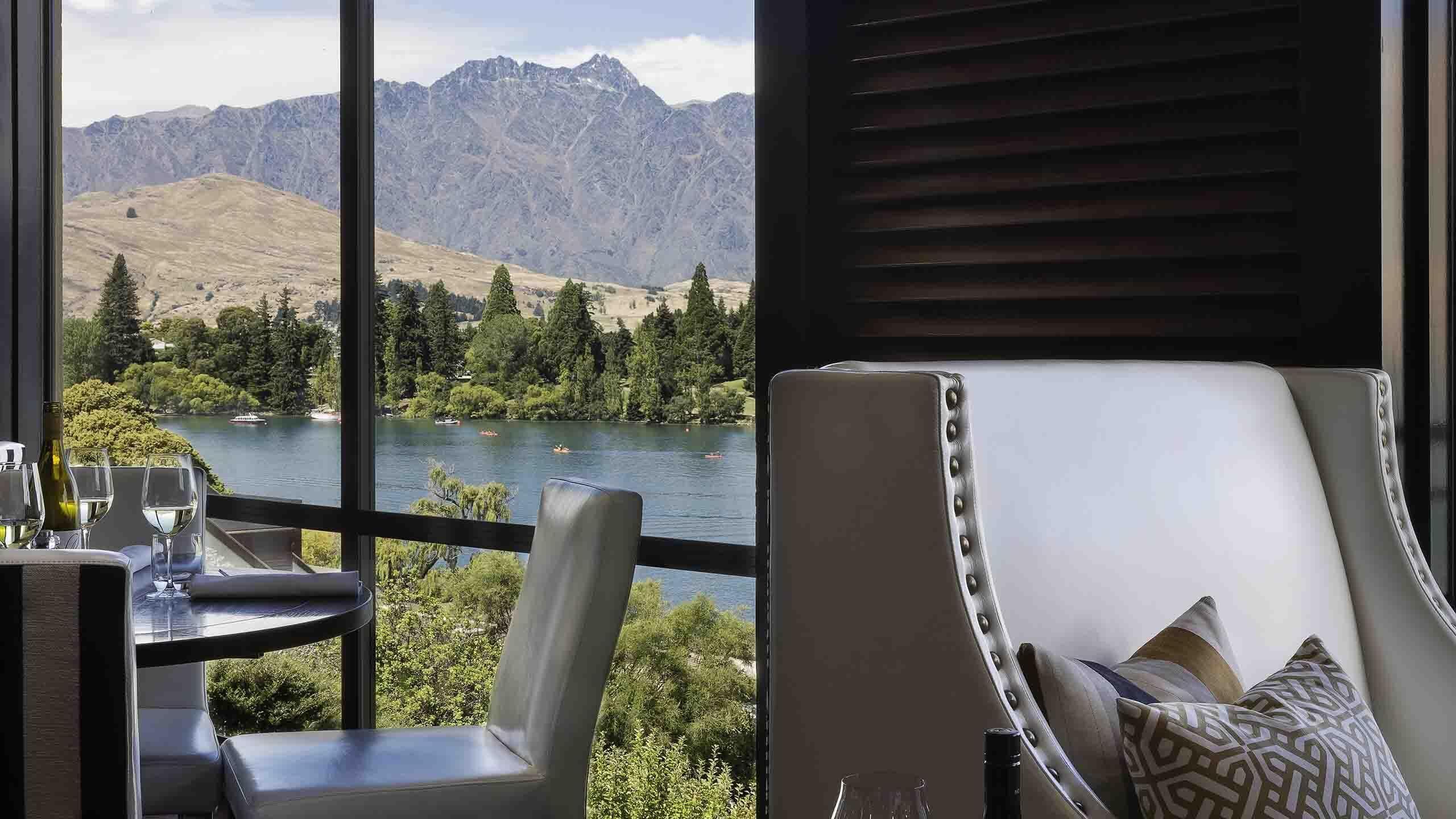 Un comedor con una mesa lista para dos personas, con vista a un lago que tiene barcos y montañas al fondo.