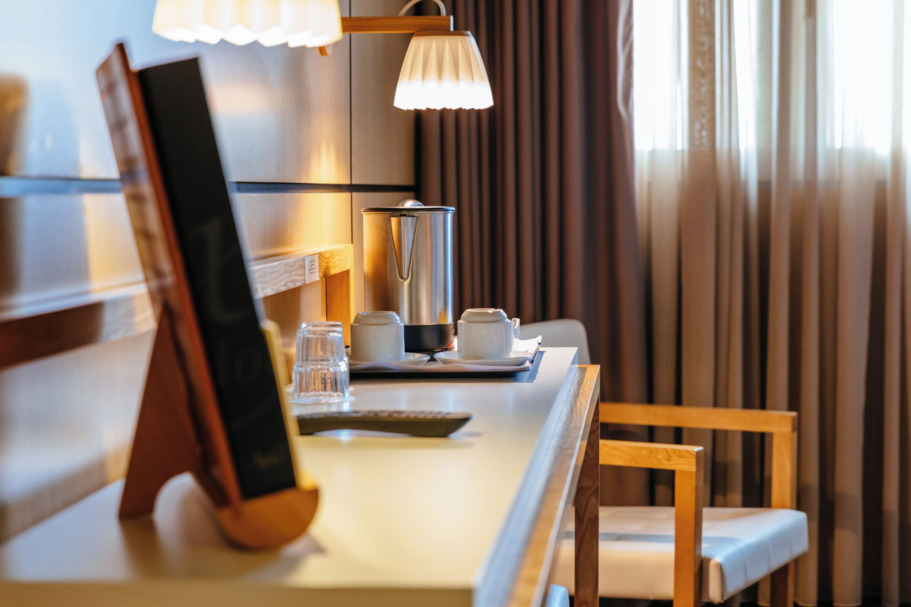A hotel room with a desk, a lamp, a coffee maker, and a chair.