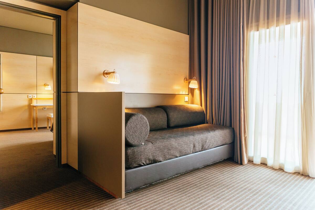 A modern hotel room with a sofa bed, two wall-mounted lamps, a window with curtains, and a visible door leading to another room.
