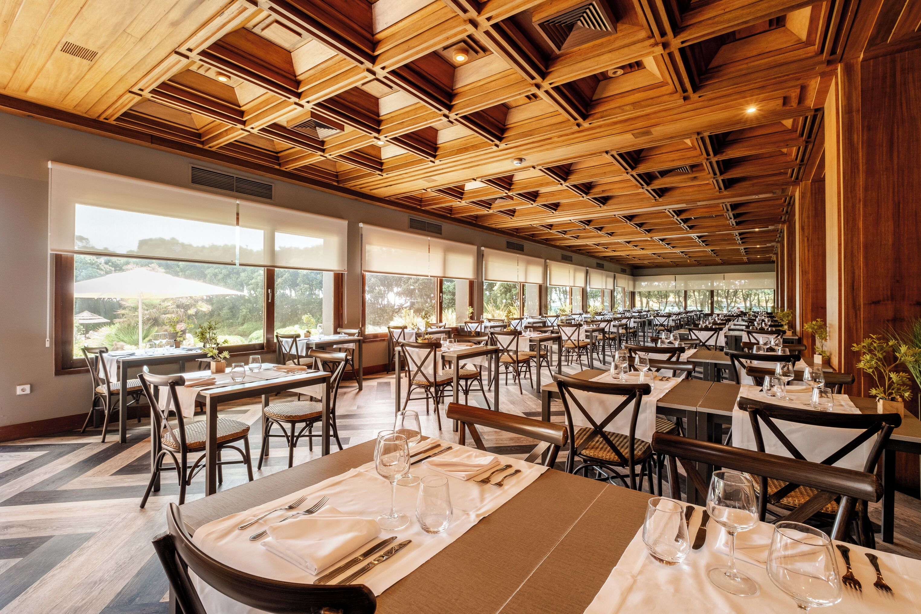 A spacious dining area with multiple tables and chairs, large windows offering views of the outdoors, and a wooden ceiling with geometric patterns.