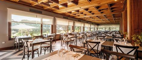 A spacious dining area with multiple tables and chairs, large windows offering views of the outdoors, and a wooden ceiling with geometric patterns.