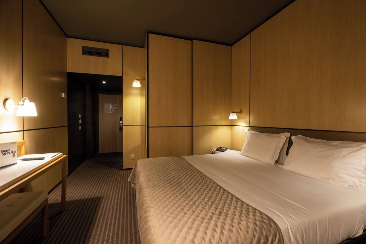 A hotel room with a bed, bedside table, lamp, and a desk with a chair.