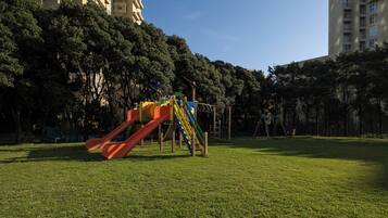 A playground with a slide and climbing structures in a grassy area with trees and tall buildings in the background.
