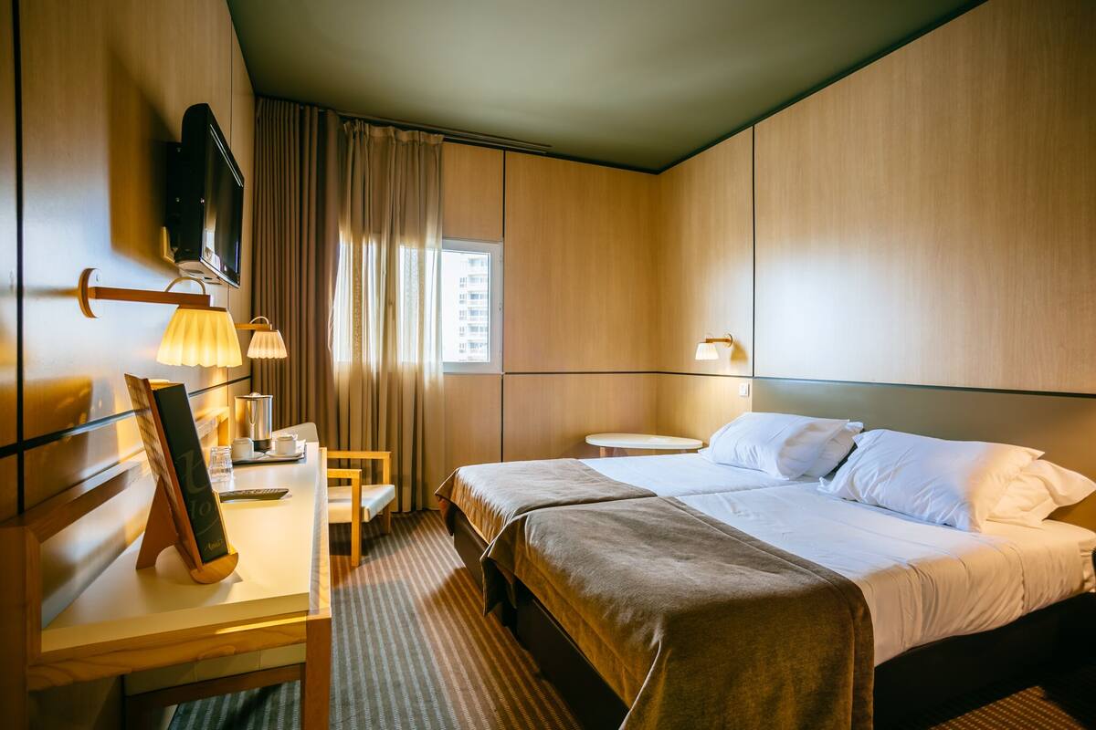 A hotel room with a bed, bedside tables, a desk with a lamp, a TV mounted on the wall, and a window with curtains.