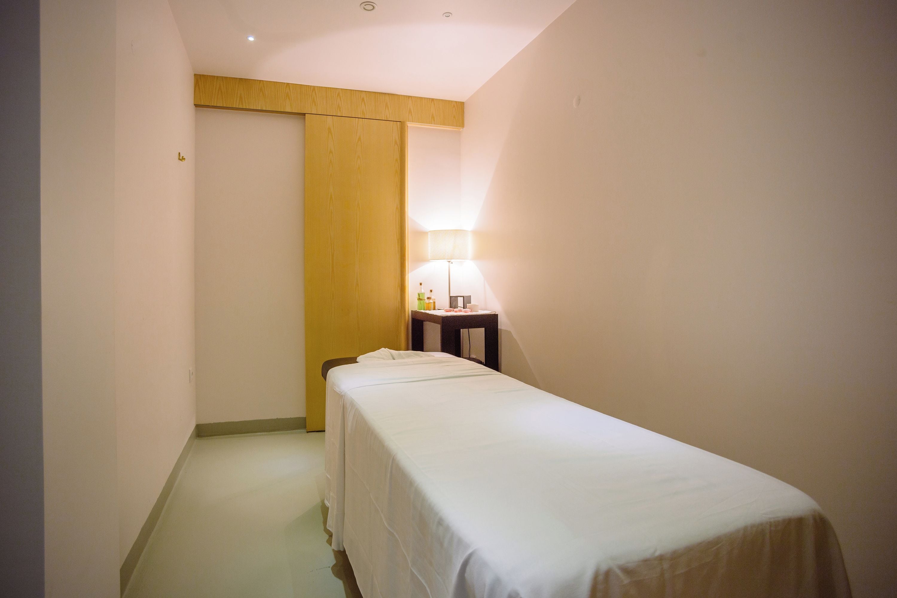 A small, narrow hotel room with a single bed, a nightstand with a lamp, and a wooden wardrobe.