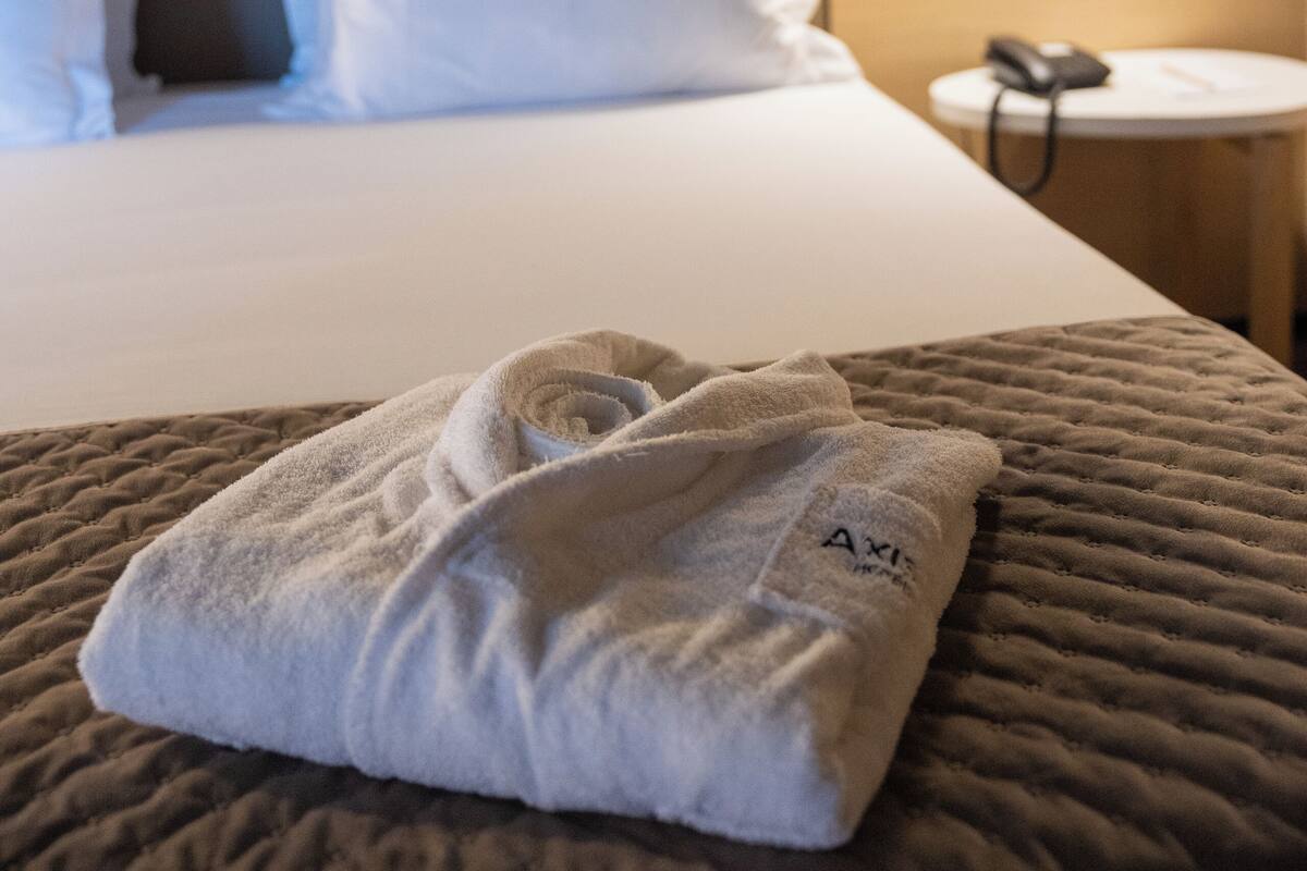 A neatly made bed with white pillows and a folded towel on a textured brown bedspread. A bedside table with a lamp and a phone is visible.