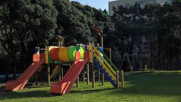 Un parque infantil con toboganes y estructuras de escalada, rodeado de árboles y un edificio al fondo.