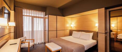 A hotel room with a bed, bedside table, chair, desk, and a view of the outside through the window.