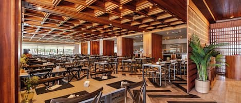 Um restaurante amplo com vigas de madeira no teto, várias mesas e cadeiras, e uma grande planta em vaso.