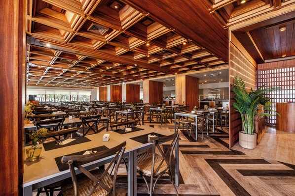 A spacious restaurant with wooden ceiling beams, multiple tables and chairs, and a large potted plant.
