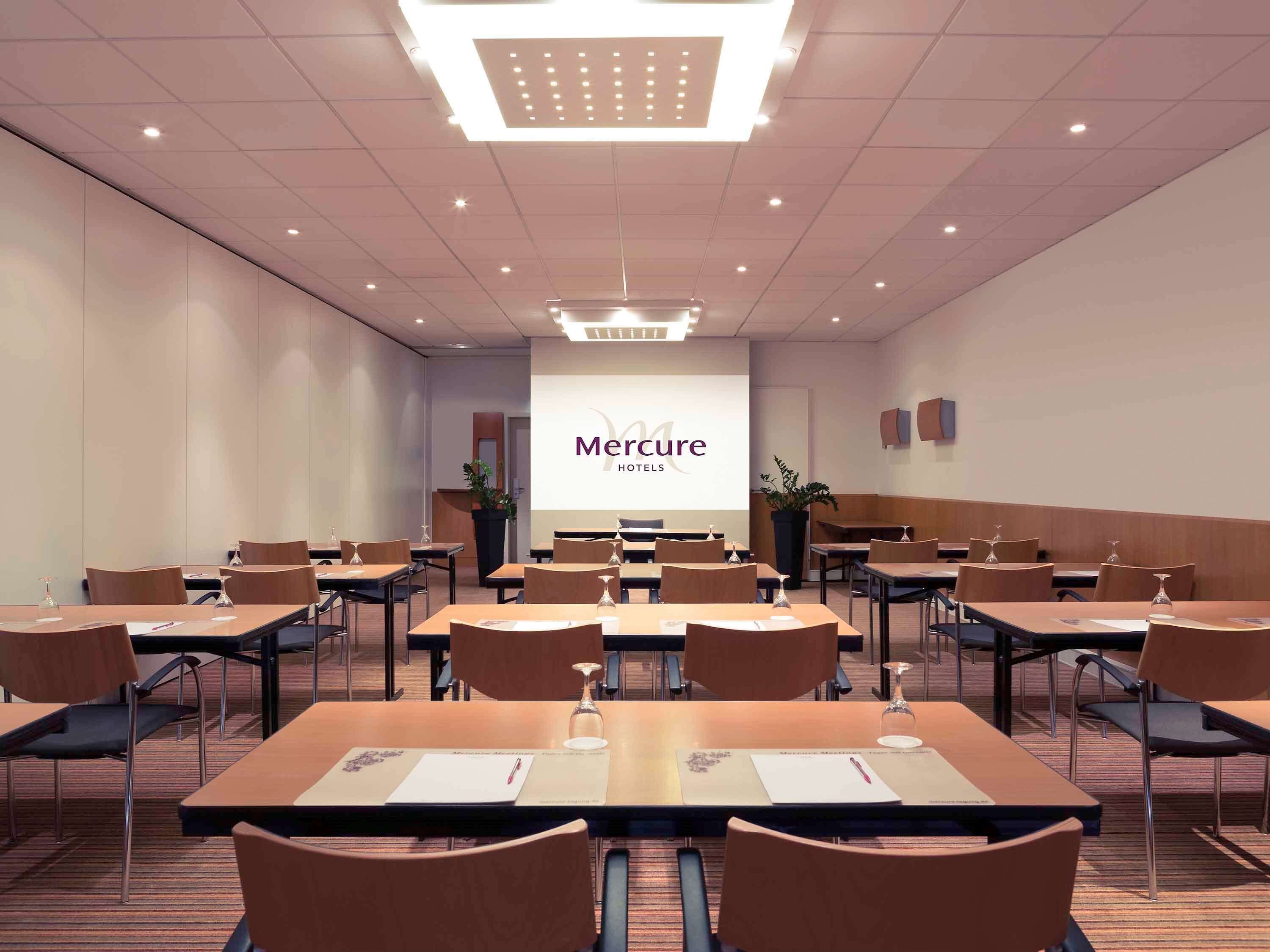 A conference room with multiple tables and chairs, a large screen displaying 'Mercure Hotels', and a modern ceiling design.