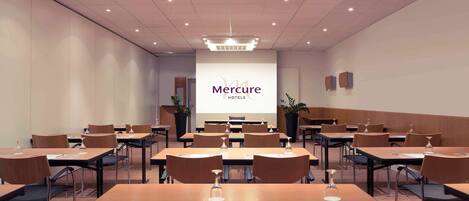 Un salón de conferencias con varias mesas y sillas, una pantalla grande que muestra 'Mercure Hotels', y un diseño de techo moderno.
