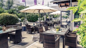 An outdoor patio area with wicker furniture and umbrellas, surrounded by greenery and a modern building in the background. - Mercure Hotel Stuttgart Sindelfingen an der Messe (Sindelfingen)