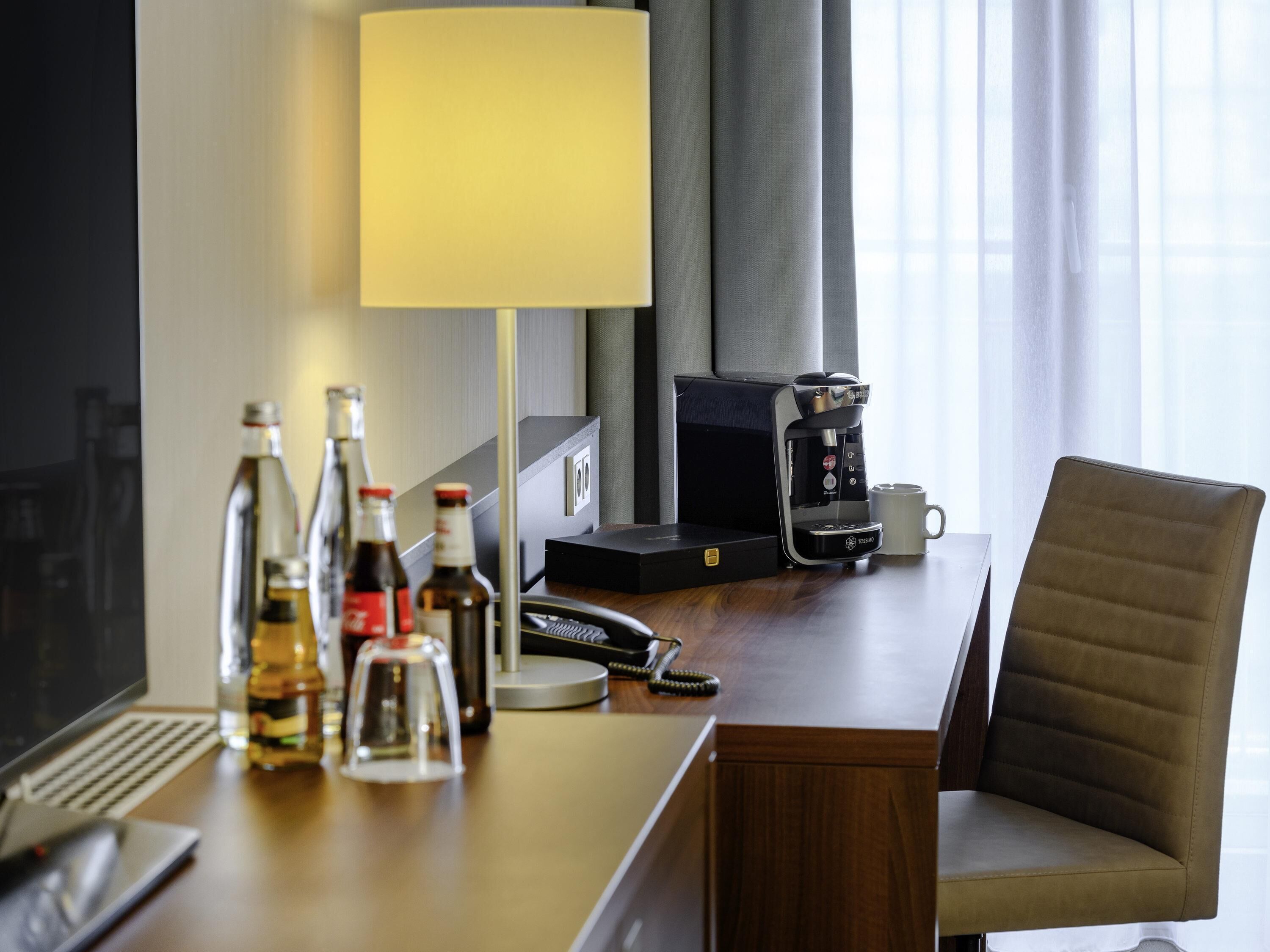A hotel room with a desk, a coffee maker, a lamp, a chair, and a computer.