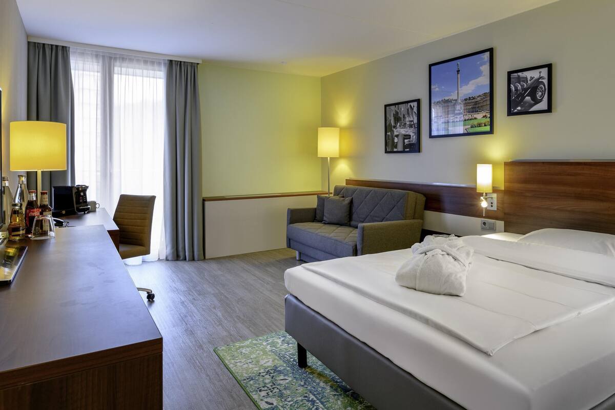A hotel room with a bed, a desk with a lamp, a chair, a sofa, and a coffee machine.