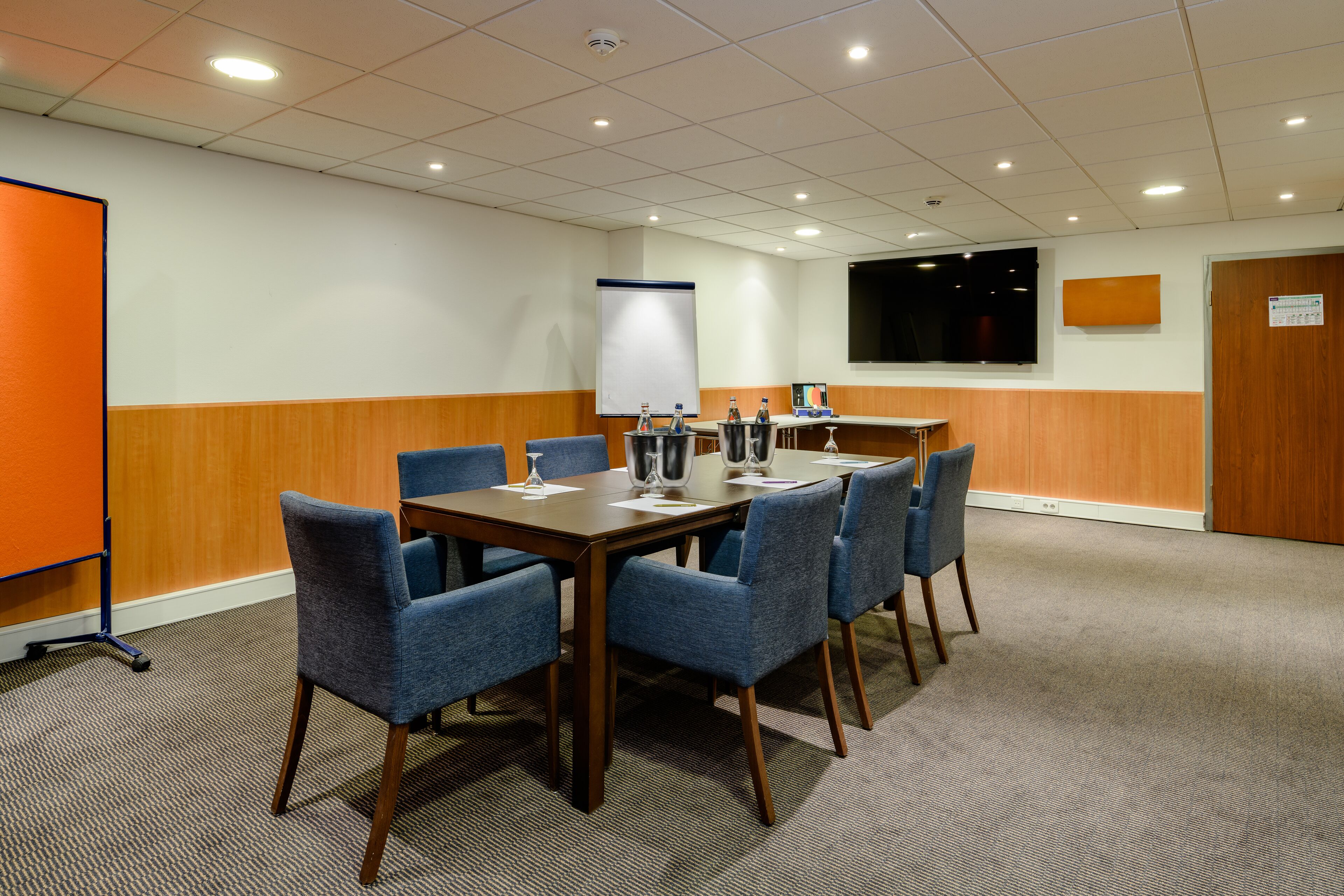 A conference room with a large table, six chairs, a whiteboard, a projector screen, and a wooden accent wall.