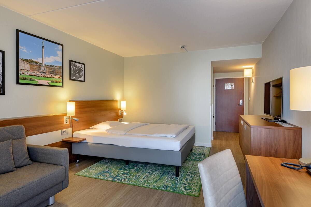 A hotel room with a bed, a desk, a chair, a sofa, and a TV.