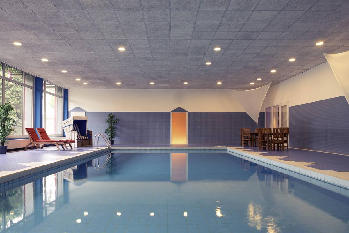 An indoor swimming pool with clear blue water, surrounded by lounge chairs, a small table, and potted plants.