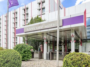 Welcome to Mercure Hotel, where a beautiful entrance is sheltered by a transparent canopy and surrounded by lush greenery.