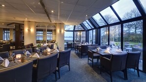 Un restaurante con techos altos, ventanas amplias y vistas al exterior. Las mesas están dispuestas con servilletas blancas y copas.