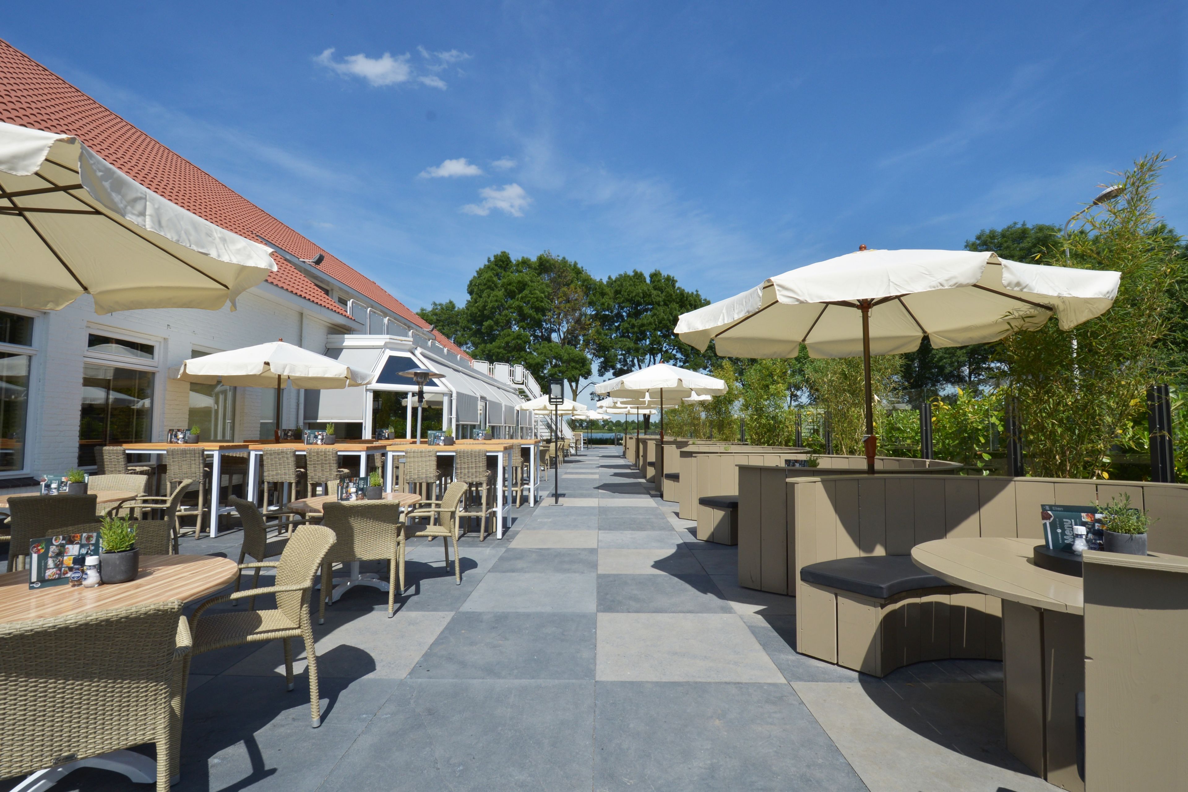 Un espace extérieur aménagé en terrasse, avec plusieurs tables et chaises, dont certaines sont dotées de parasols, et un chemin menant à un bâtiment dont le toit est rouge.