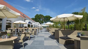 Un espace extérieur aménagé en terrasse, avec plusieurs tables et chaises, dont certaines sont dotées de parasols, et un chemin menant à un bâtiment dont le toit est rouge.