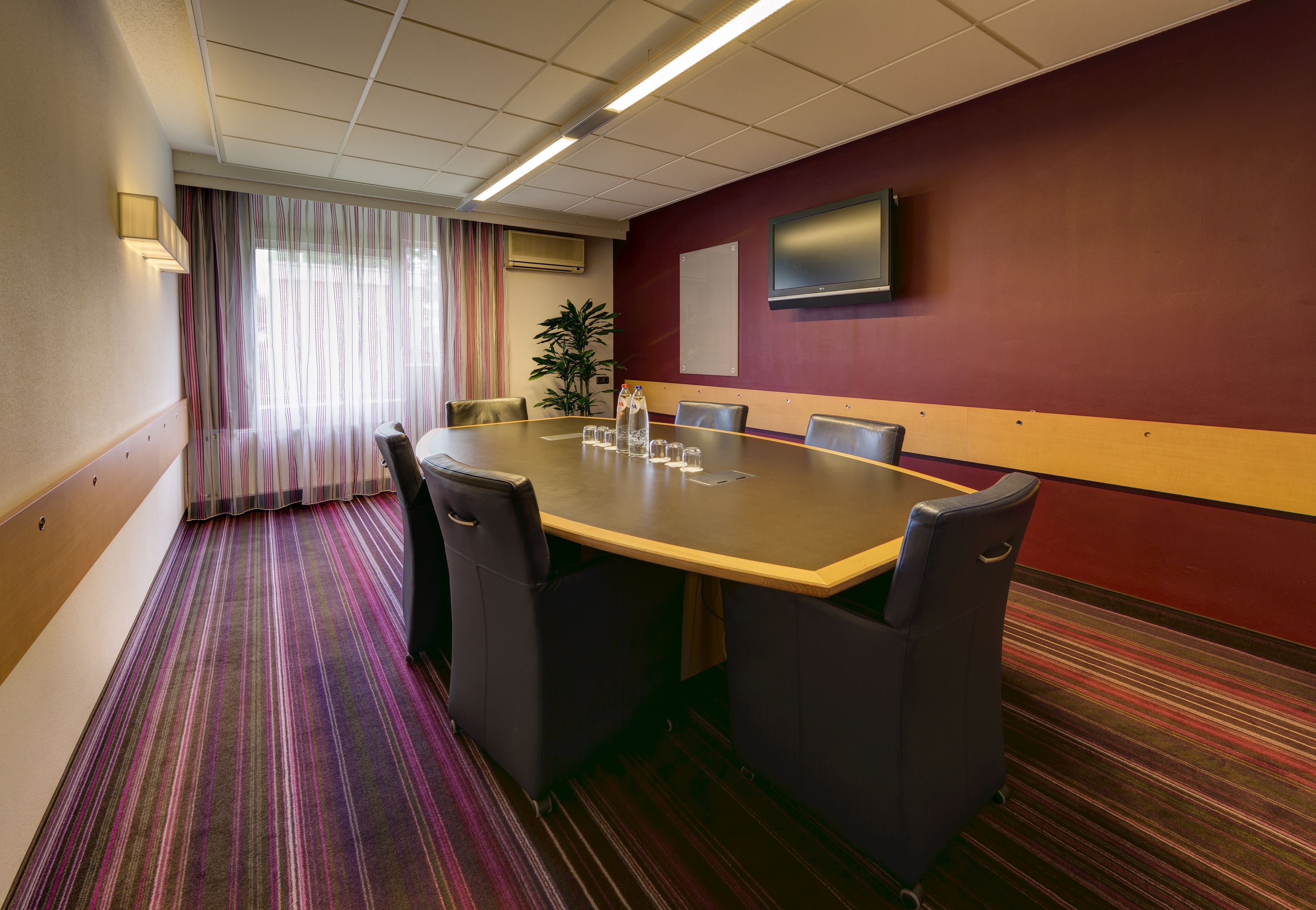 A conference room with a large table, chairs, a TV on the wall, and a striped carpet.